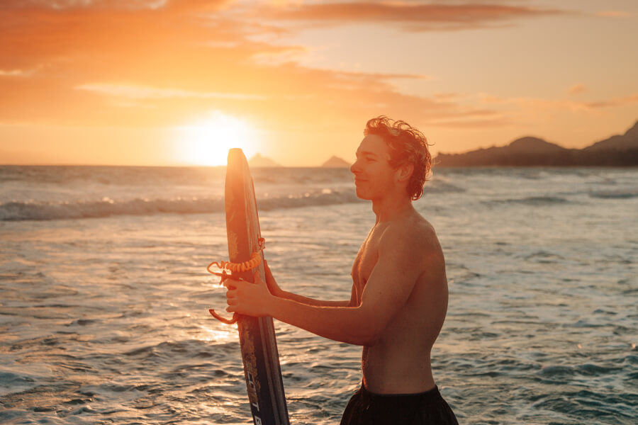 A boy holds a boogie board rental from Active Oahu while standing in the ocean water at sunset