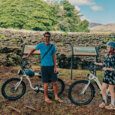 Two people standing with e-bike rentals in front of an ancient Hawaiian temple